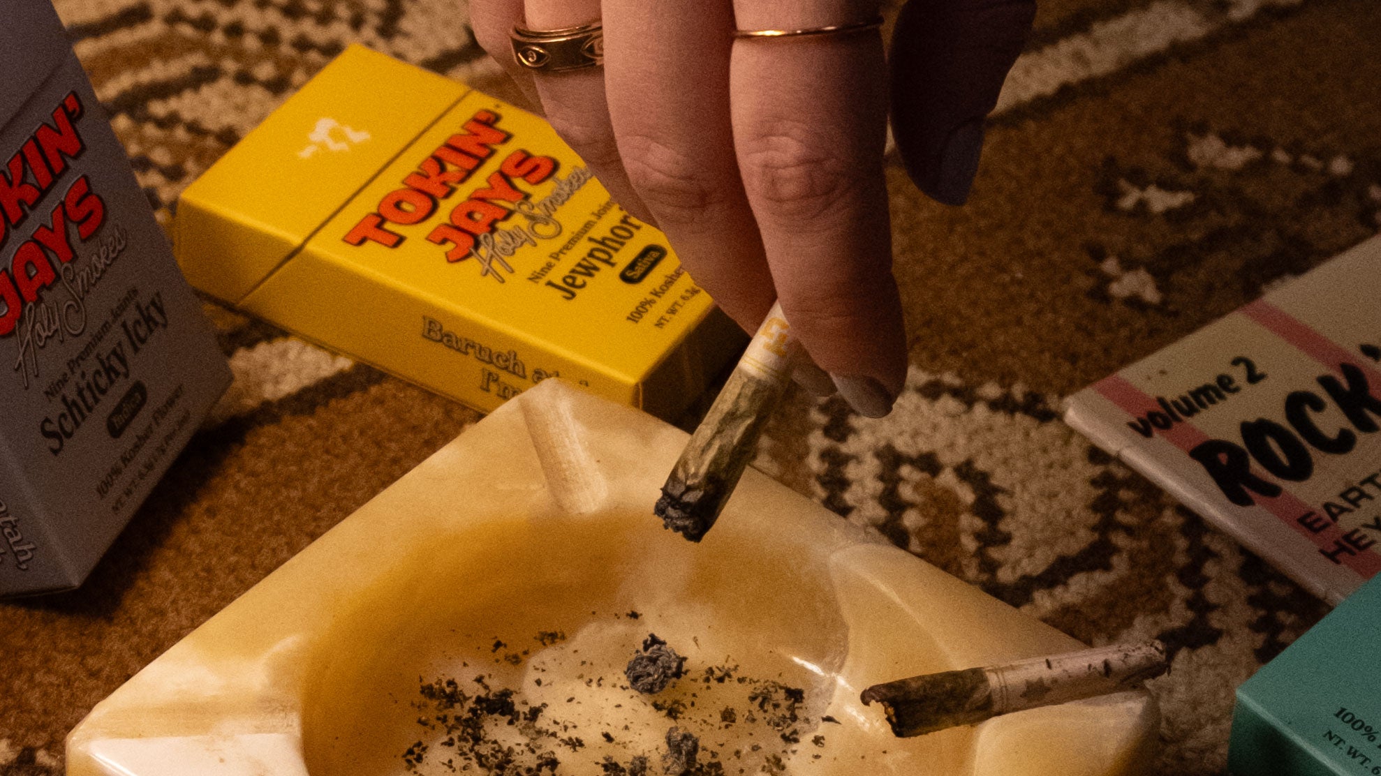 Hand holding a lit joint with a box of cigarettes and a magazine in the background.