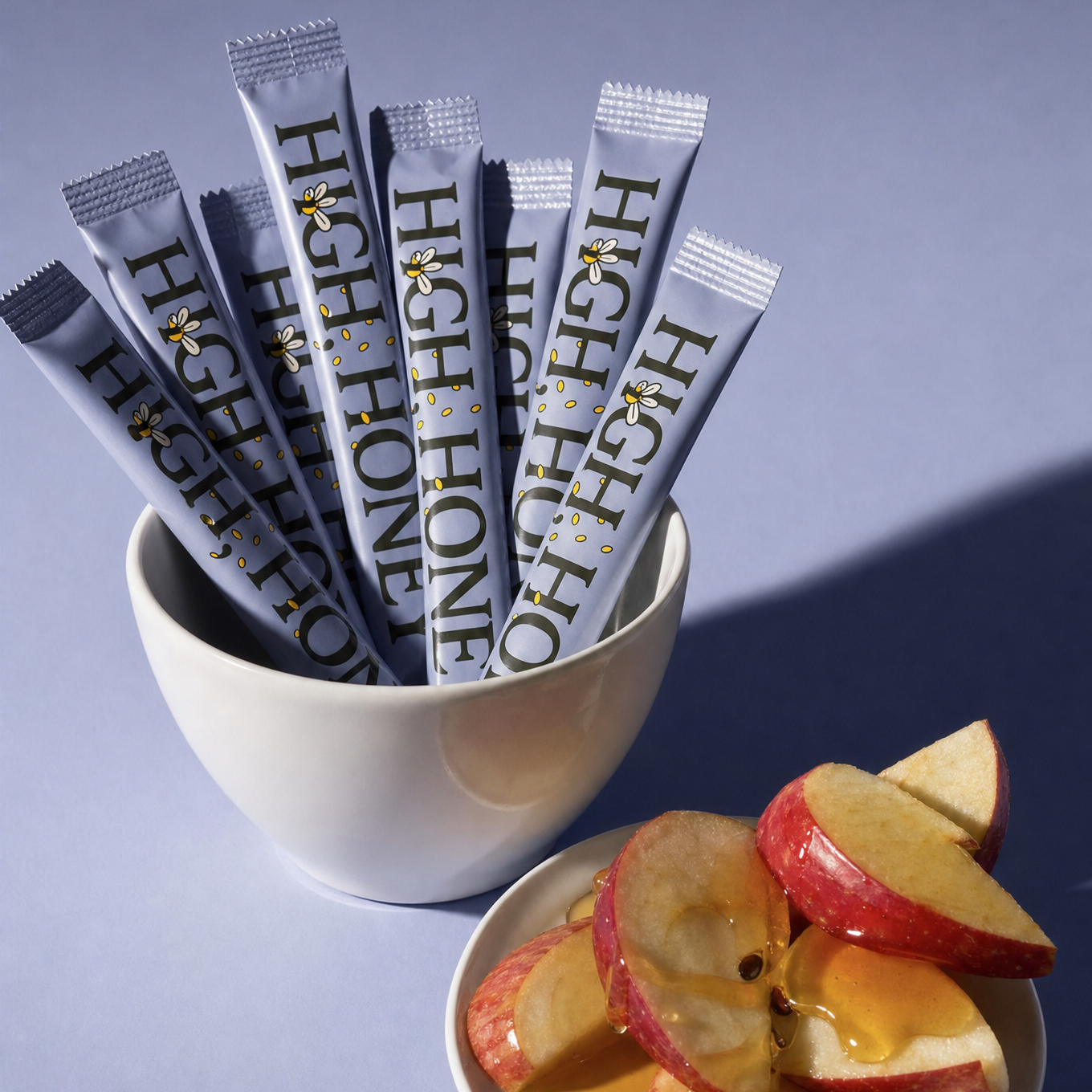 Several silver and white stick packs printed with 'HIGH HONEY' are arranged in a white bowl, with sliced red apples and honey on a small plate in front, set against a lavender-blue background.