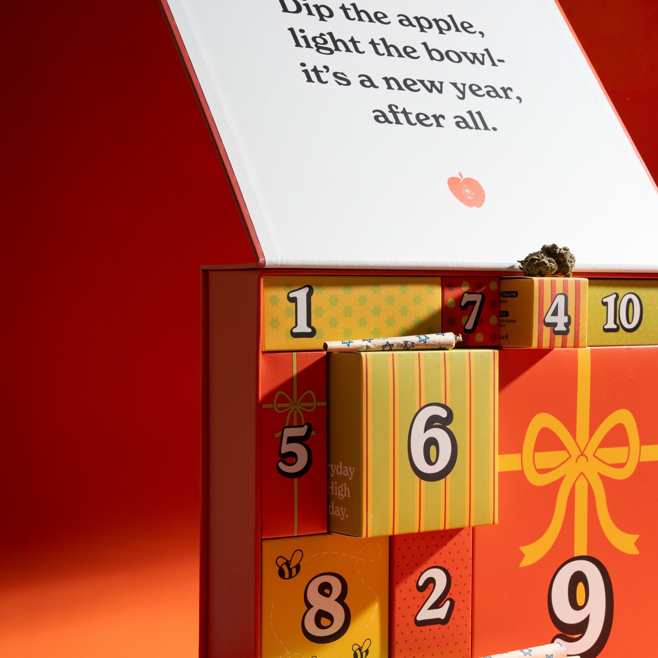 A advent calendar with numbered drawers in green, yellow, orange, and red, shown open with a white flap displaying printed text. The box is photographed on a warm orange-red studio background with strong directional light and shadows.