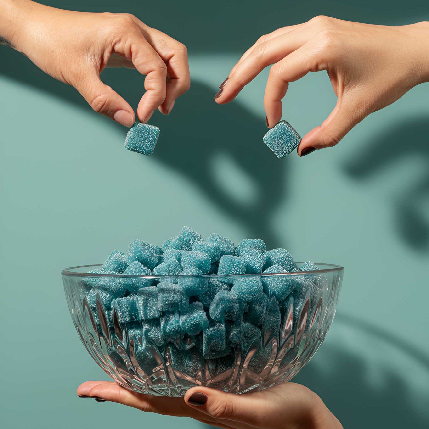 Three hands reaching towards a bowl of blue gummy cubes against a teal background