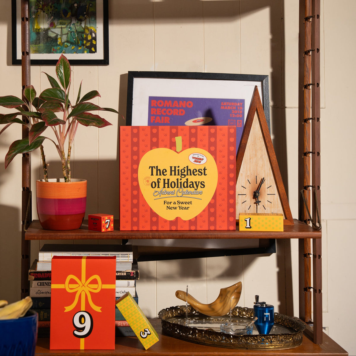 A styled holiday-themed bookshelf display featuring a red and yellow 'The Holidays' book, colorful wooden number blocks, a wooden clock, framed art, a potted plant, and gift boxes on multiple shelves.