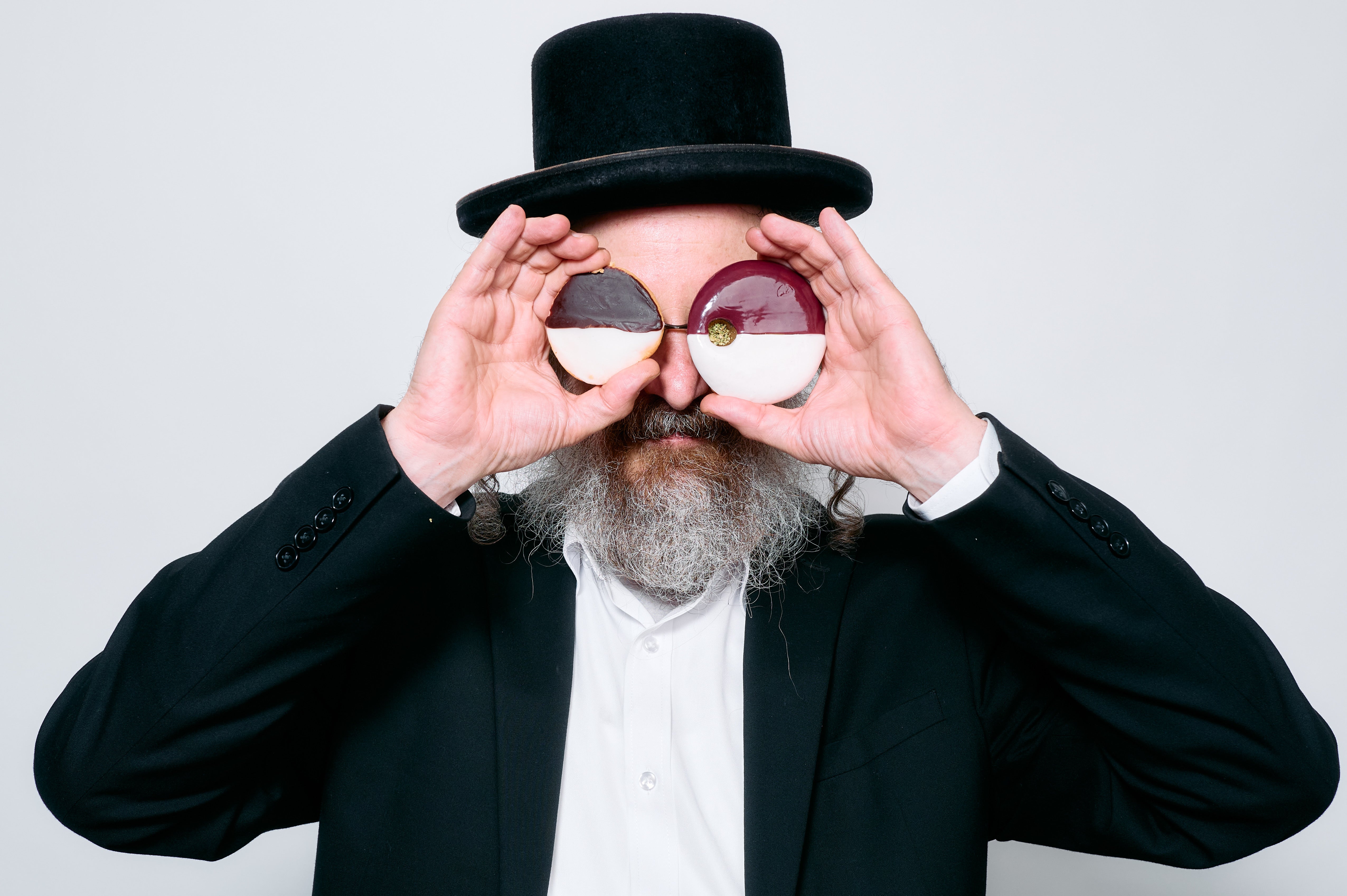 A man with a gray beard wearing a black shtreimel, black suit jacket, and white shirt, holding a pair of black & white cookies and a pipe over his eyes like binoculars against a light gray background.