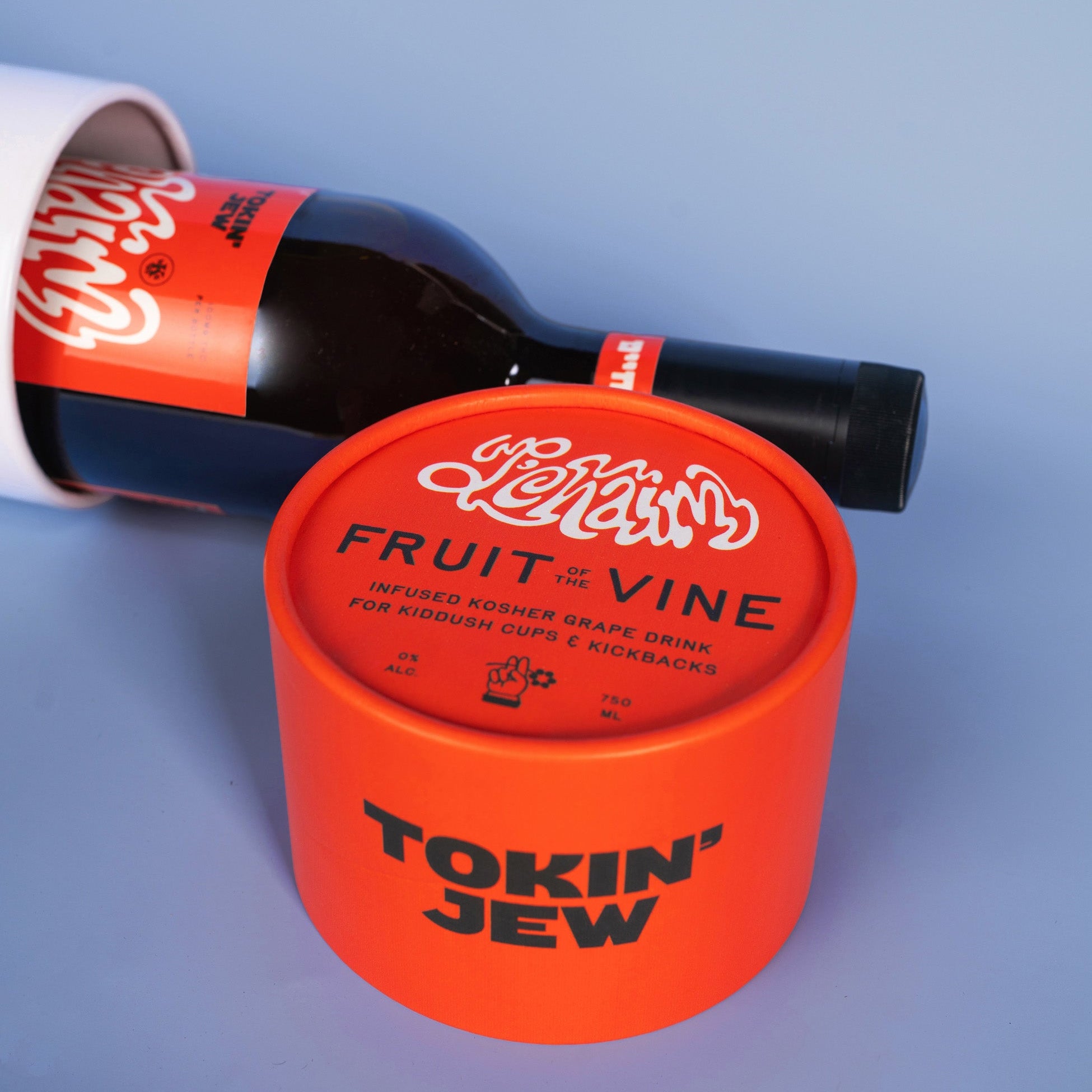 Tokin' Jew infused grape drink packaging, featuring a bright orange cylindrical tub in the foreground with the brand name 'TOKIN' JEW' and product name 'Fruit of the Vine', and a dark glass bottle with an orange label lying behind it on a light blue background.