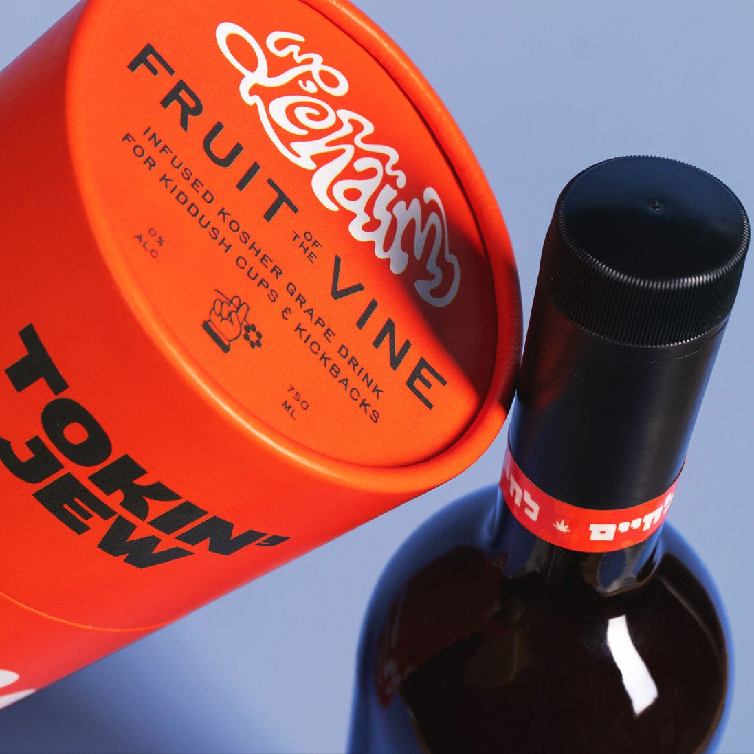 An orange cylindrical product container labeled 'Fruit of the Vine' and 'Tokin' Jew' lies diagonally next to a dark glass bottle with a black cap on a light blue background.
