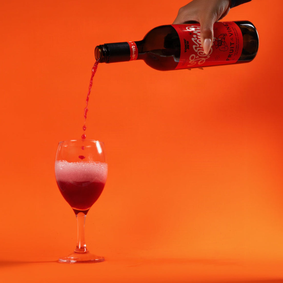 A hand holds a bottle of red juice with a black cap and red label, pouring the red liquid into a stemmed glass against a solid bright orange background.