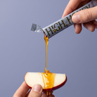 A hand holds a foil packet of High Honey syrup above a red apple slice, with golden honey dripping from the packet onto the apple against a solid blue-gray background.