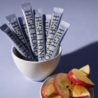 Several silver and white stick packs printed with 'HIGH HONEY' are arranged in a white bowl, with sliced red apples and honey on a small plate in front, set against a lavender-blue background.