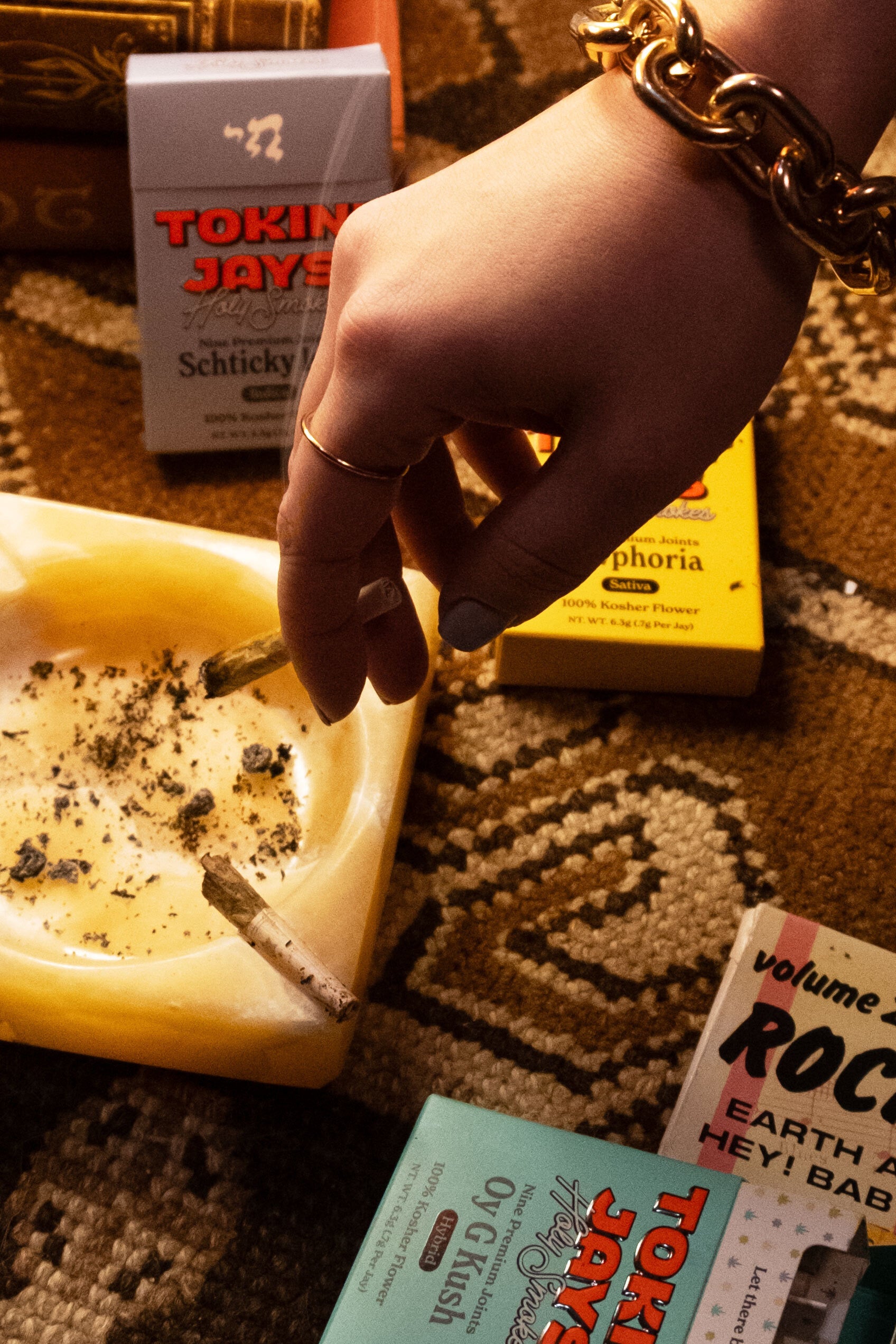 hand tipping a tokin jay joint on an ashtray, positioned on a rug with scattered tokin jays packs around it