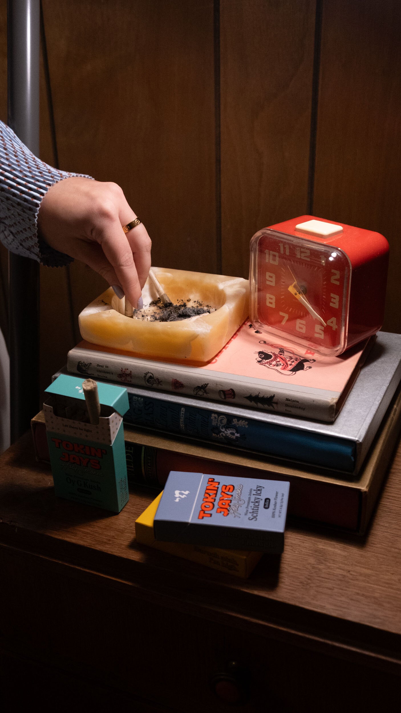 person tipping a joint in an ashtray on a bedside table with decorative books and a clock and tokin jays packs