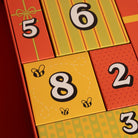 A close-up of a colorful gift advent calendar with numbered compartments. The visible sections are bright orange-red, yellow, and green with large numbers 5, 6, 8, 2, and 3 printed on the panels, plus small bee illustrations on one yellow panel.