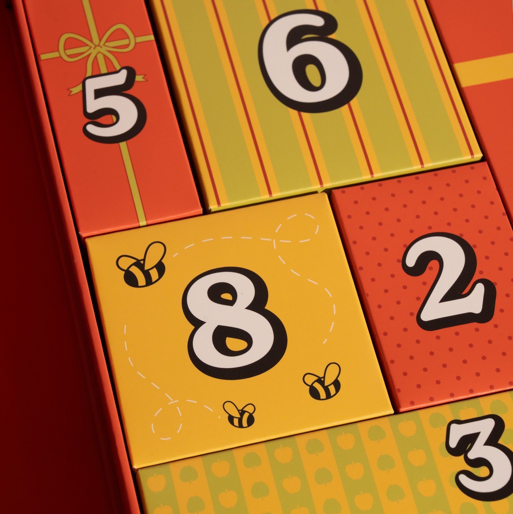 A close-up of a colorful gift advent calendar with numbered compartments. The visible sections are bright orange-red, yellow, and green with large numbers 5, 6, 8, 2, and 3 printed on the panels, plus small bee illustrations on one yellow panel.