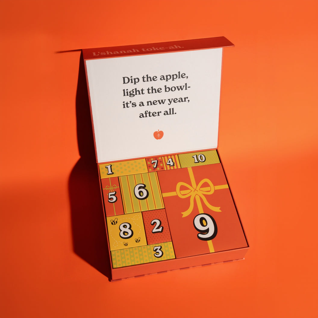 A decorative numbered advent-style box opened on a bright orange background. The inside lid has printed instructions reading, 'Dip the apple, light the bowl- it's a new year, after all.'
