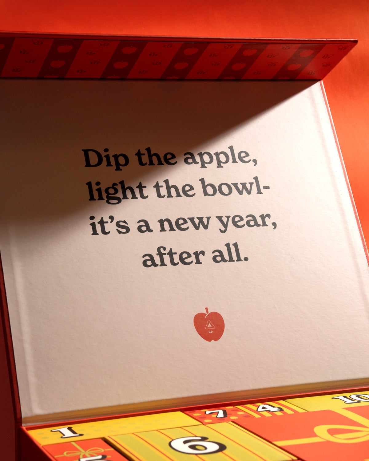 A close-up of a red and yellow decorative box with printed text reading, 'Dip the apple, light the bowl- it's a new year, after all.' and a small red apple icon near the bottom.