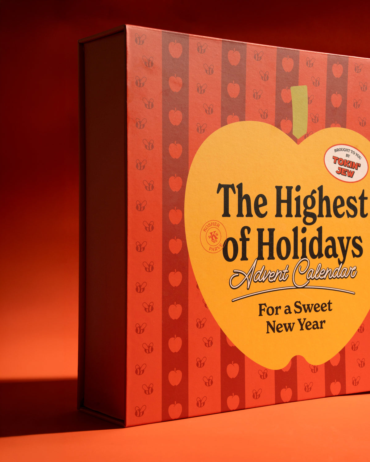 A boxed holiday advent calendar titled "The Highest of Holidays Advent Calendar For a Sweet New Year" shown in a warm orange-red studio setting. The box is decorated with vertical red and orange stripes and a large yellow apple-shaped panel on the front.