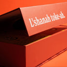 A close-up of a red-orange product box with the Hebrew text 'L'shanah toke-ah.' printed in white on the lid, set against a matching red-orange background.