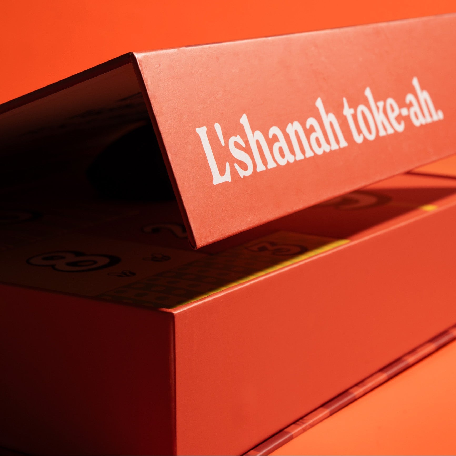 A close-up of a red-orange product box with the Hebrew text 'L'shanah toke-ah.' printed in white on the lid, set against a matching red-orange background.