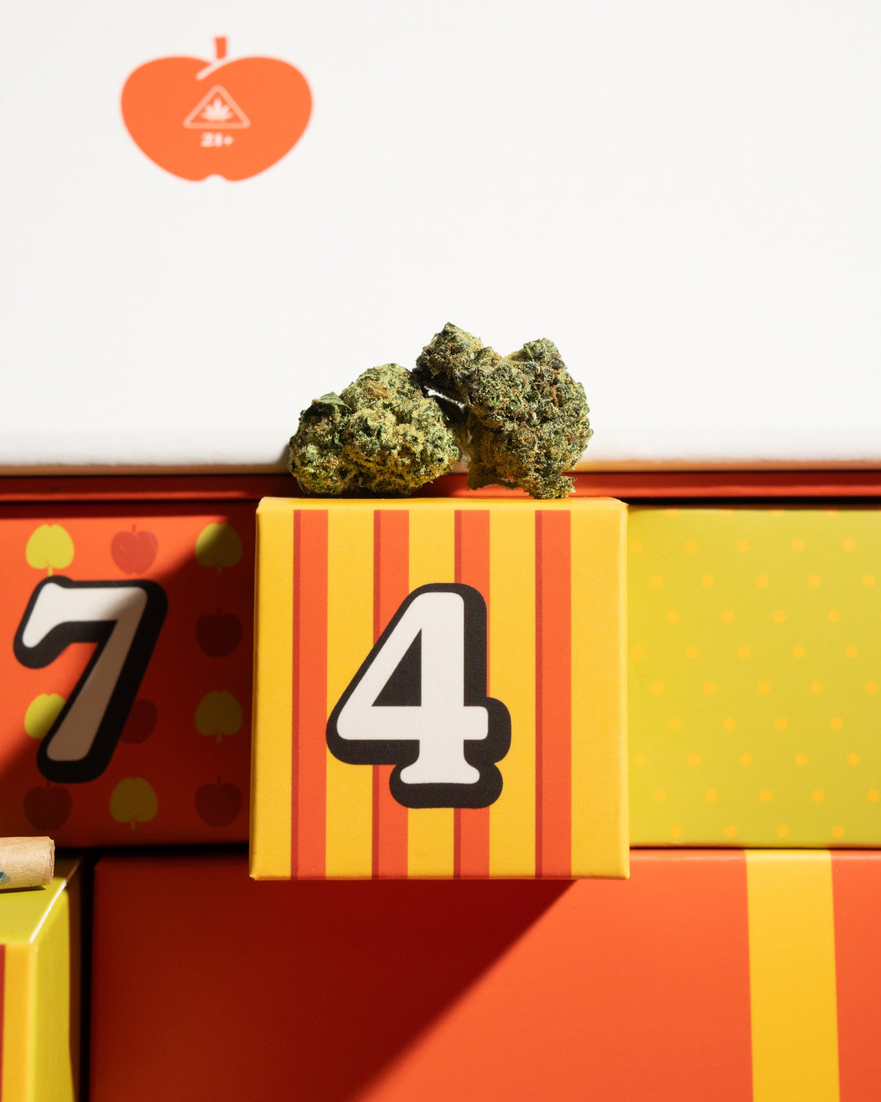 A close-up of colorful product packaging with numbered boxes, including a yellow box marked '4' and another marked '7', with dried green buds resting on top. An orange apple-shaped logo with a cannabis leaf is visible in the upper left against a white background.