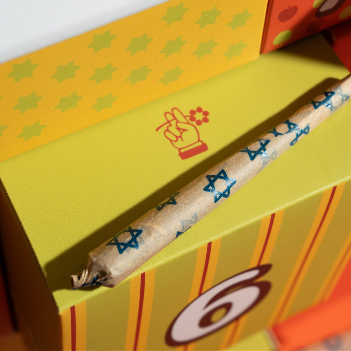 A close-up of a rolled joint wrapped in patterned paper resting on a yellow and orange striped box with a hand graphic printed on the lid.