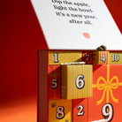 A advent calendar with numbered drawers in green, yellow, orange, and red, shown open with a white flap displaying printed text. The box is photographed on a warm orange-red studio background with strong directional light and shadows.