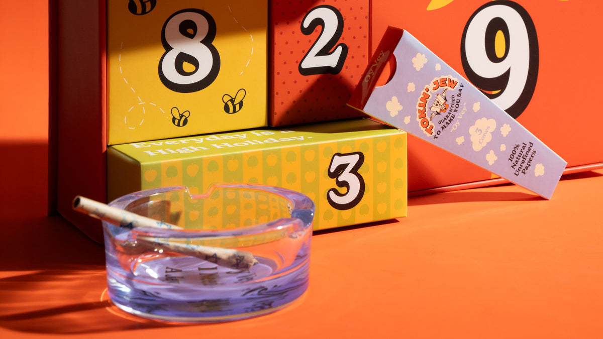 A styled product shot featuring a clear glass ashtray with a lit joint resting inside, positioned in front of colorful numbered boxes (8, 2, 3) and a lavender branded packet on a bright orange background.