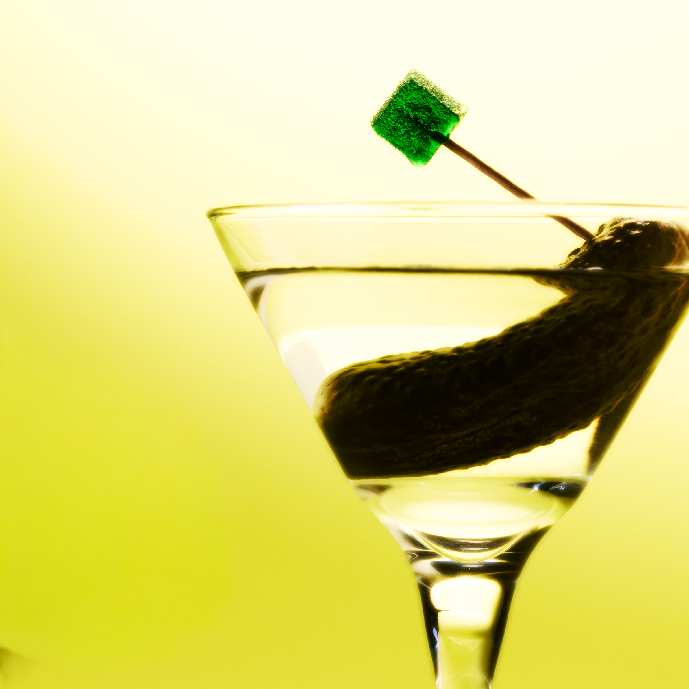 Martini glass with a pickle gummy on a toothpick against a yellow background