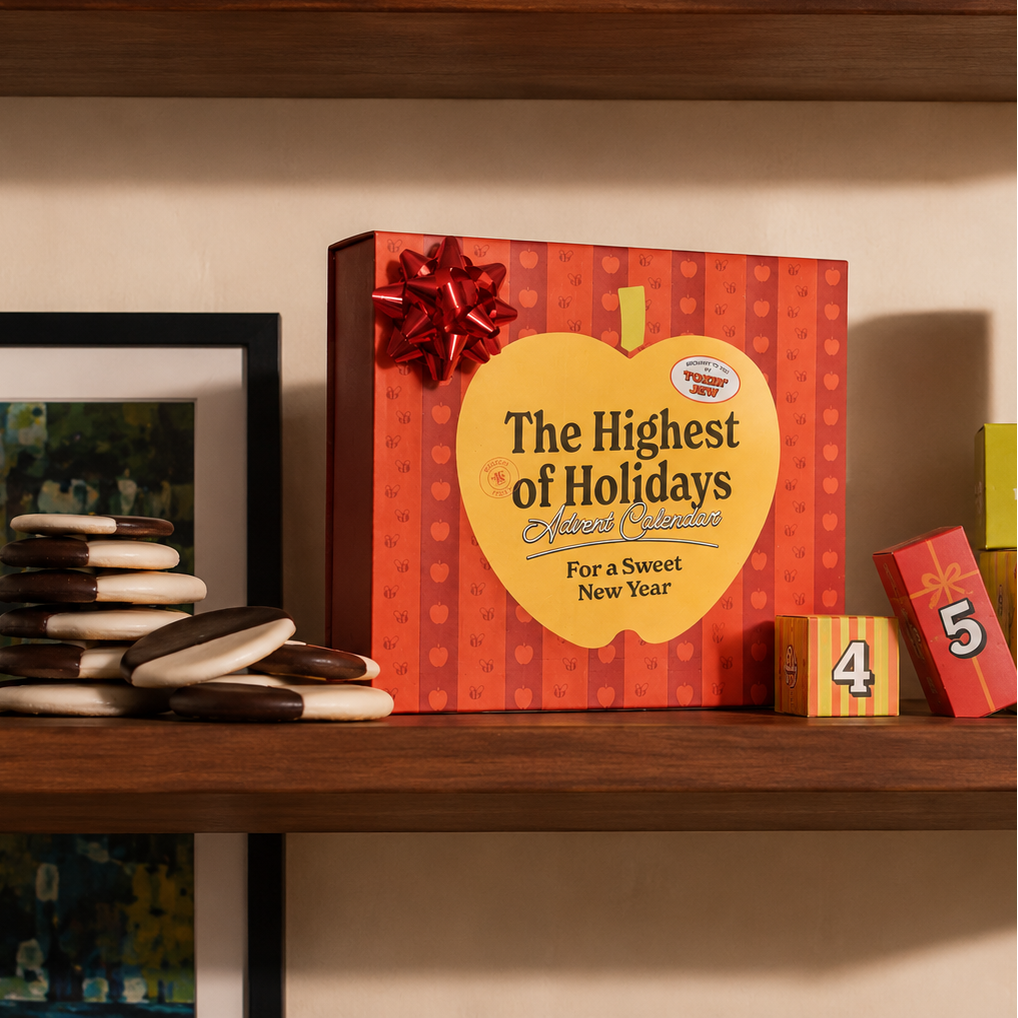 A boxed holiday gift set labeled "The Highest of Holidays" sits on a wooden shelf beside stacked chocolate sandwich cookies, framed artwork, and colorful numbered blocks. The box is red with an apple-shaped front panel and a red bow.