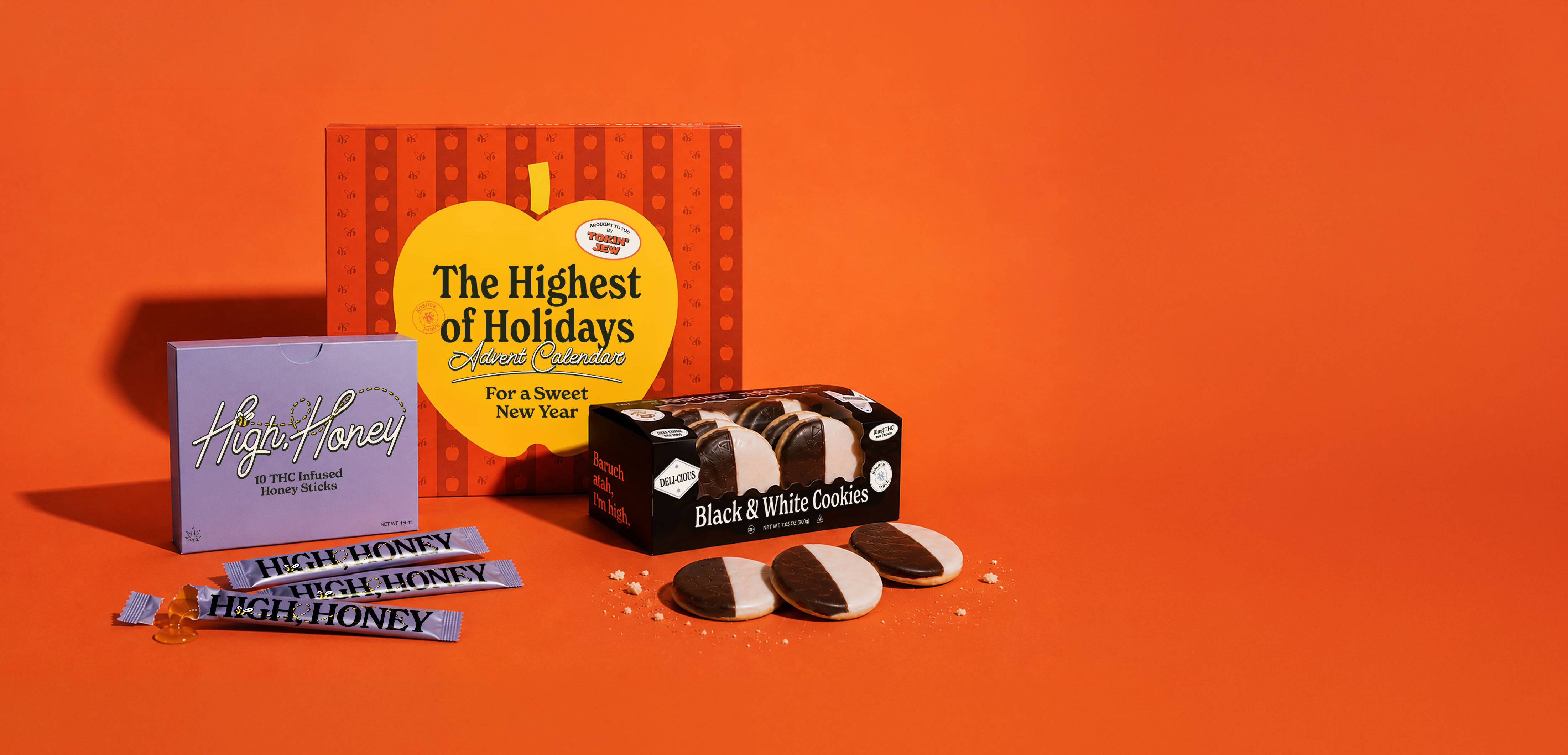 A holiday-themed snack and cookie gift set arranged on a bright orange background. The set includes a purple box labeled 'High Honey' with several blue-topped honey sticks, a black box of 'Black & White Cookies', and a red-and-yellow hanging ornament-shaped box reading 'The Highest of Holidays Sweet New Year'.