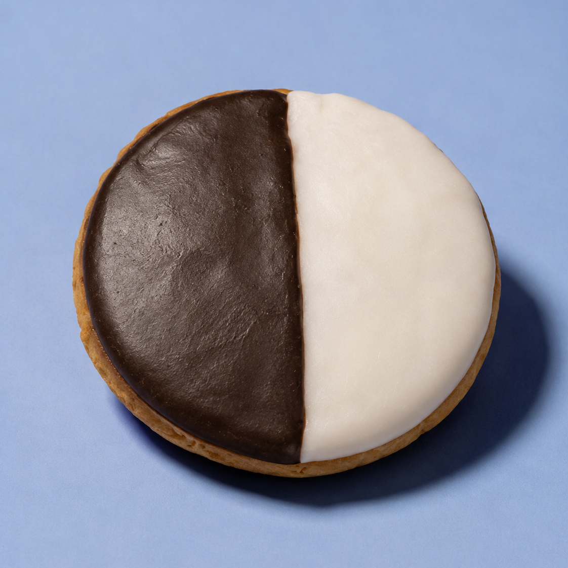 A round iced cookie split vertically into two halves: one half dark brown and one half white, placed on a smooth light blue background.
