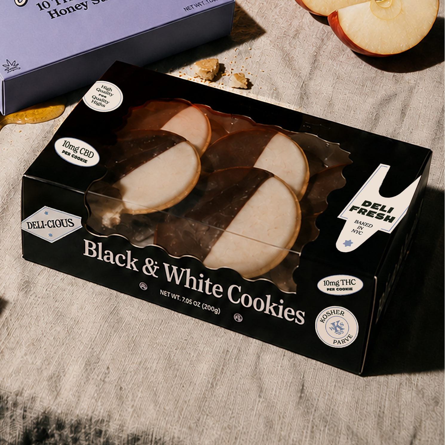Boxed gourmet treats arranged on a draped fabric surface with apple slices, crumbs, and honey props: a purple box labeled 'High Honey' and a black box labeled 'Black & White Cookies,'.