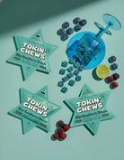 Star-shaped 'Tokin' Chews' packages with blue raspberry flavor on a light green background with berries and a blue shaker.