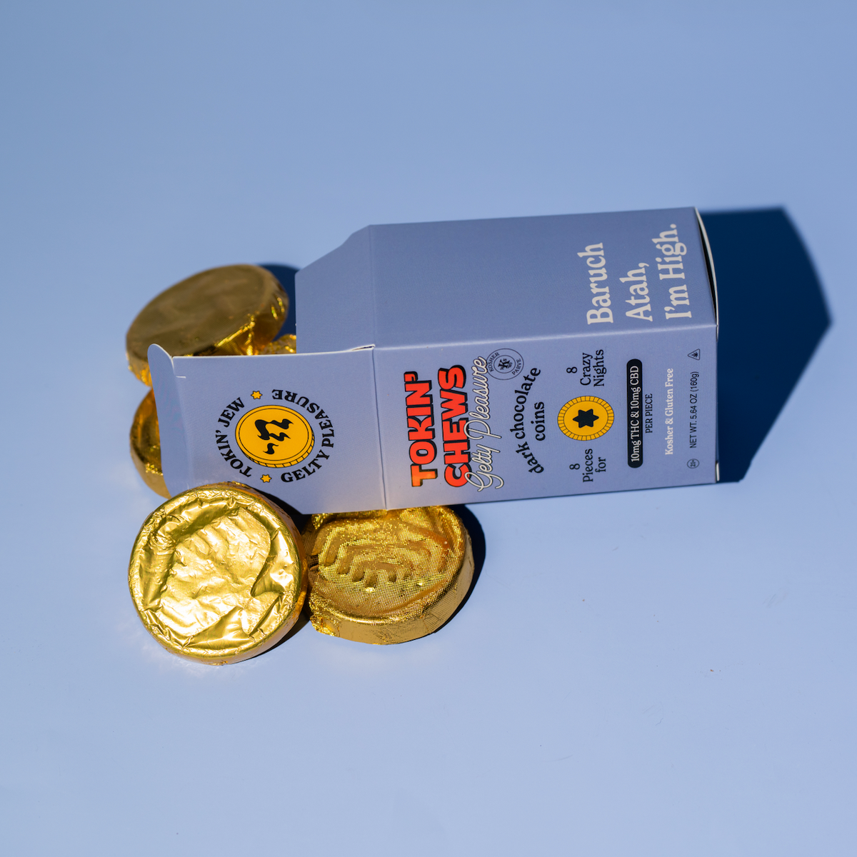 A small silver and white box labeled 'Tokin Chews' with several gold foil-wrapped coin pieces scattered in front, photographed on a smooth light blue background.