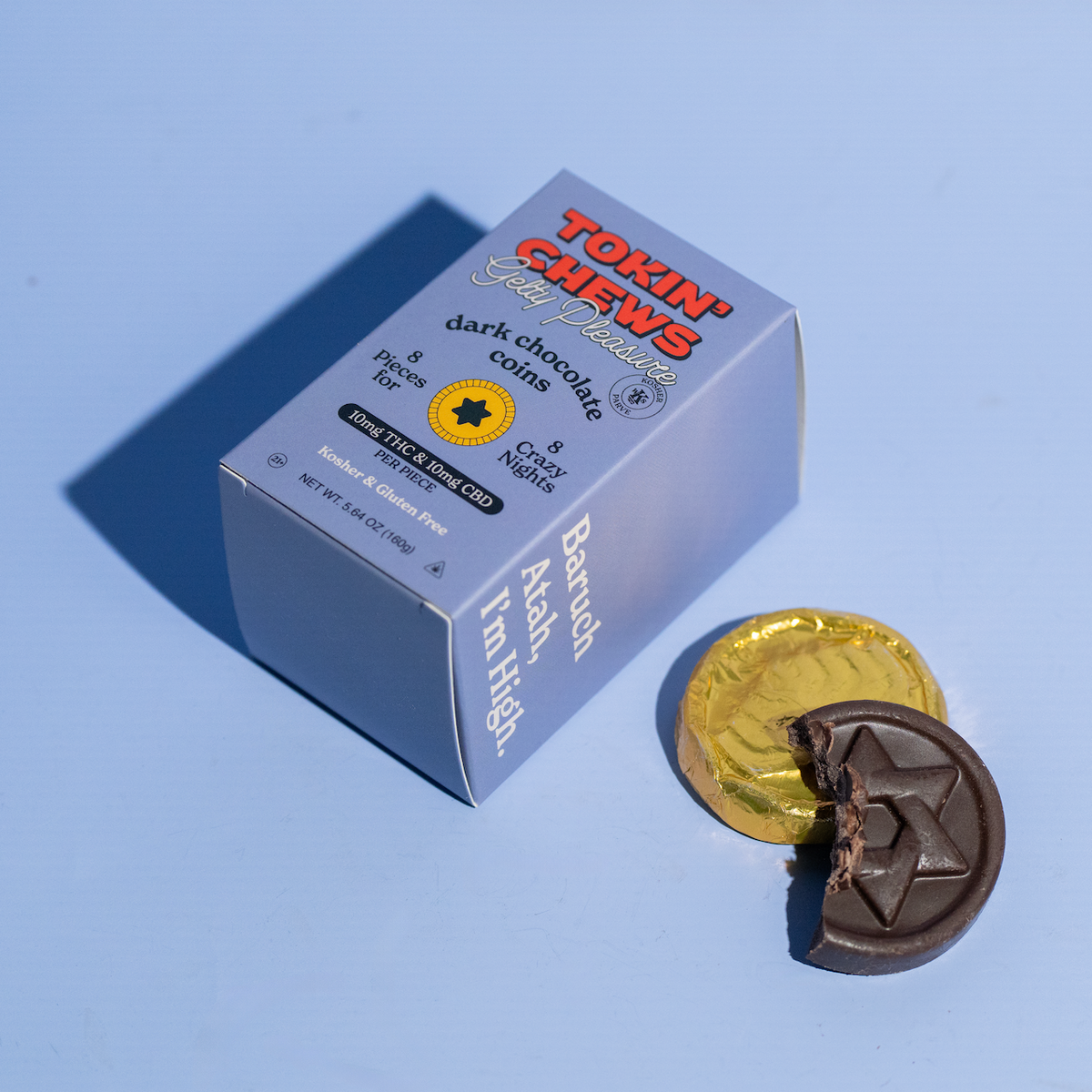 A box of Tokin' Chews dark chocolate gummies alongside two chocolate coins, one of which is broken to show the star-shaped center, set against a solid light blue background.