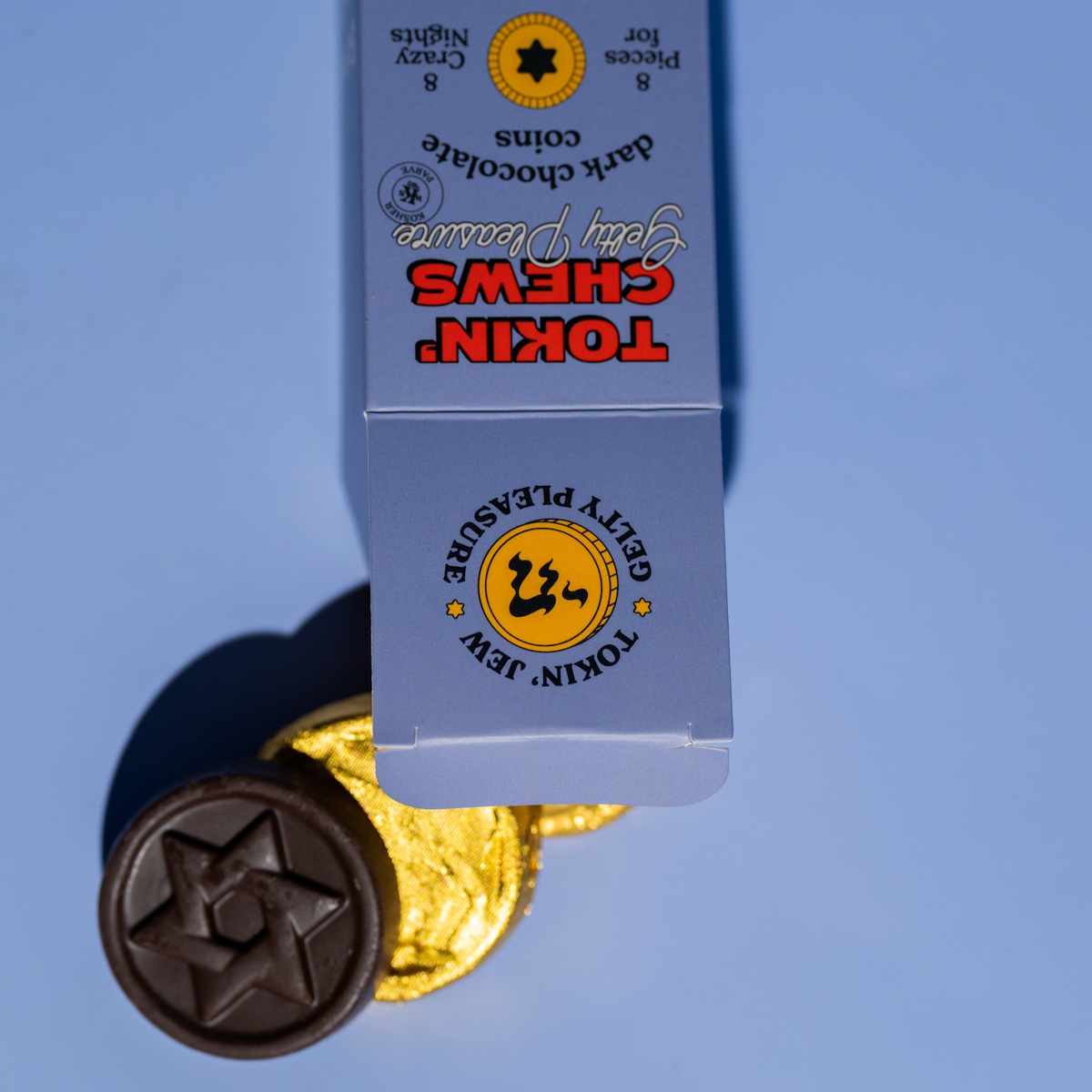 A boxed set of Tokin' Chews dark chocolate coins and a single wrapped chocolate coin on a light blue background. The packaging is light blue with red and yellow branding, and the chocolate coin is wrapped in gold foil with a dark brown center.