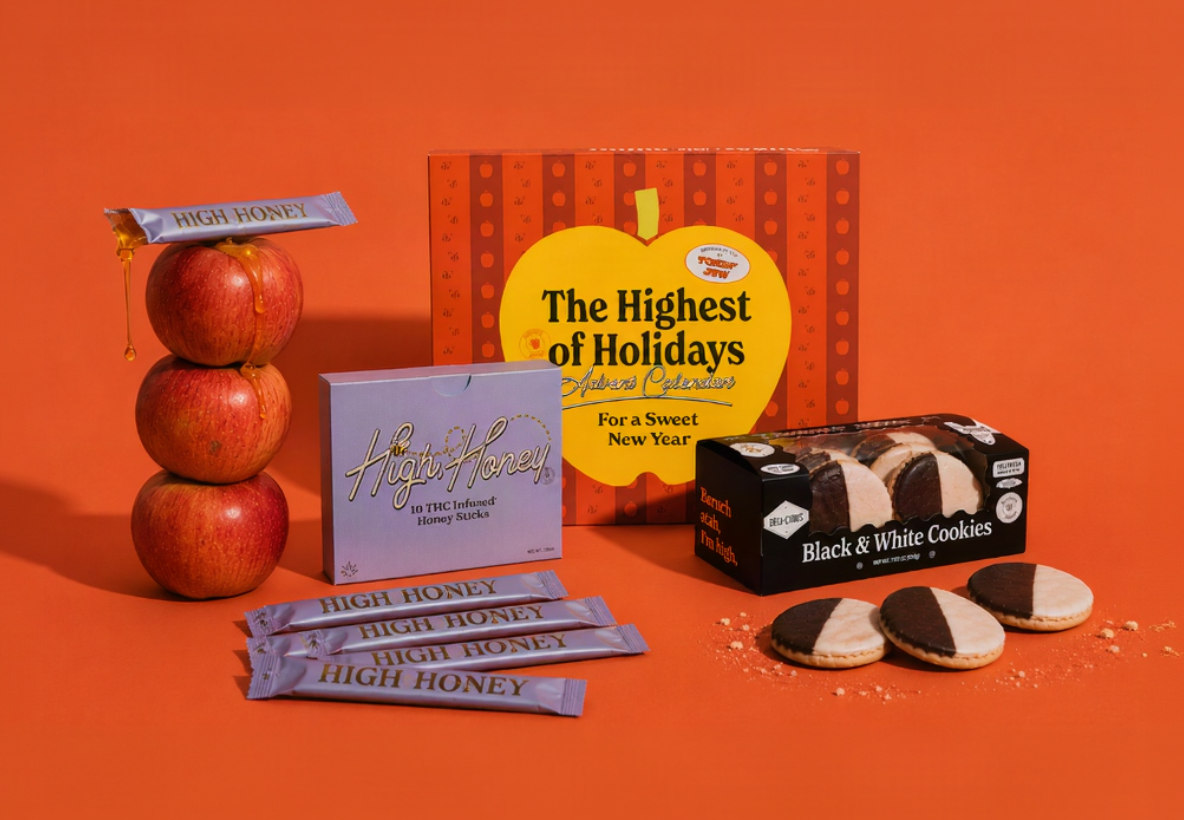 A styled product display featuring High Honey products, including stacked red apples with a honey stick on top, several individual high honey sticks, a lavender box of honeycomb, a box of black and white cookies, and a large orange and yellow apple-shaped box reading 'The Highest of Holidays Holiday Collection'.