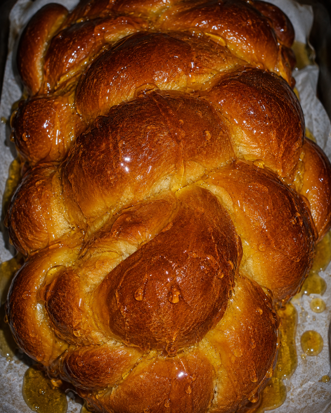 A close-up of a braided loaf of bread with a glossy golden-brown glaze, resting on parchment paper.