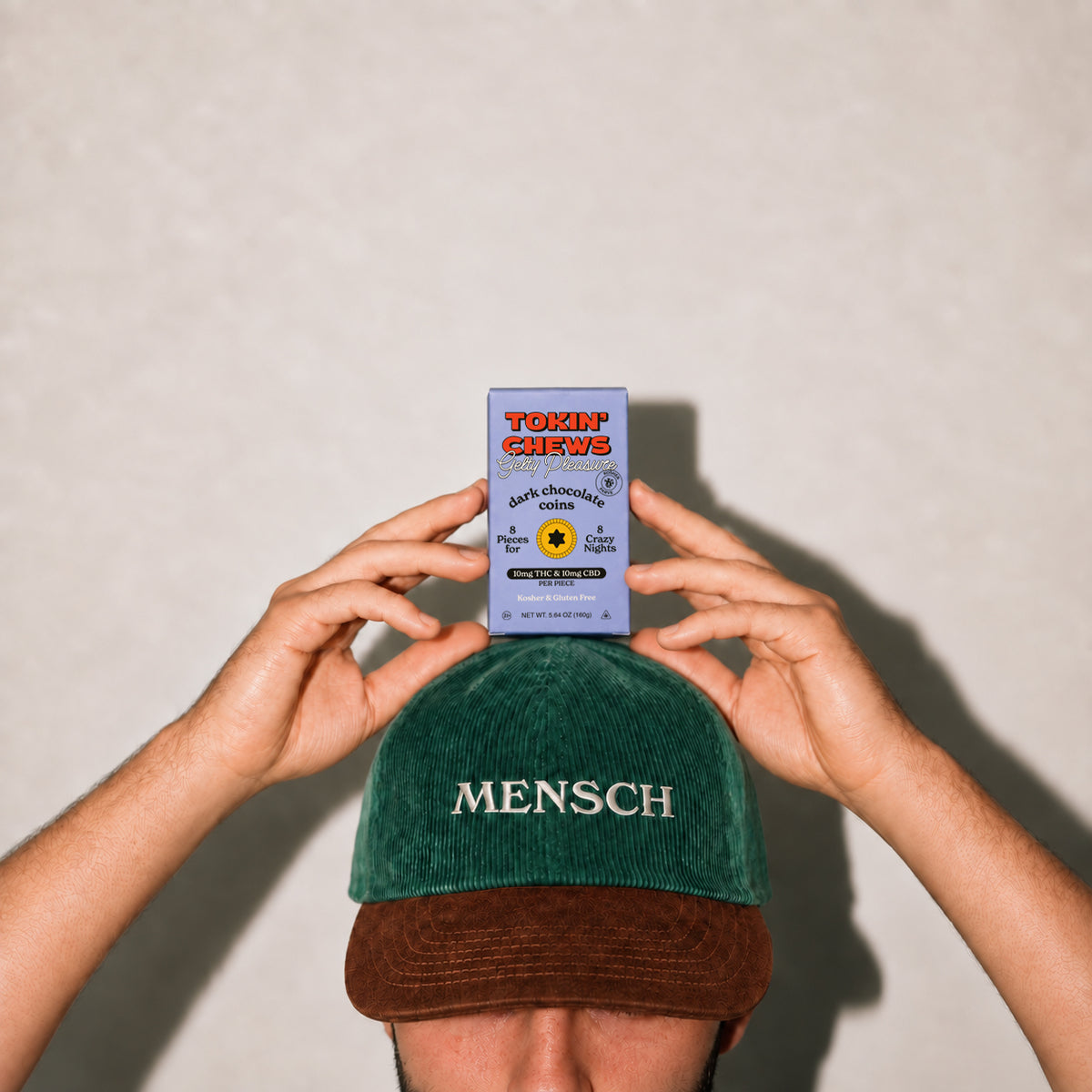 A person holds a small purple box in front of their face, wearing a green corduroy cap with 'MENSCH' embroidered in white on the front. The background is a plain light beige wall with a soft shadow.
