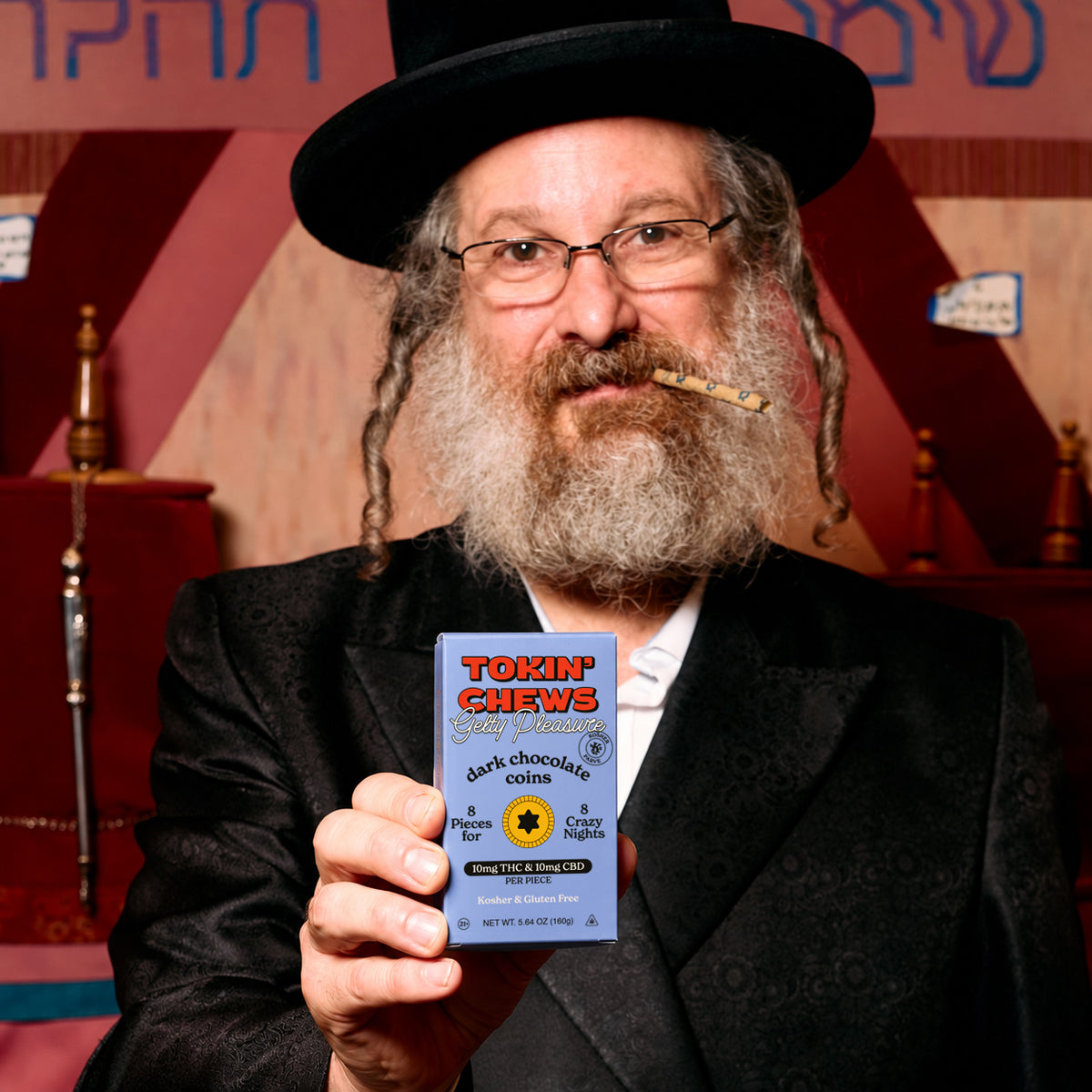 A rabbi in a black hat and suit jacket holds up a blue package of Toxin' Chews dark chocolate gelt. The background features a warm, patterned red and gold wall with Hebrew lettering.
