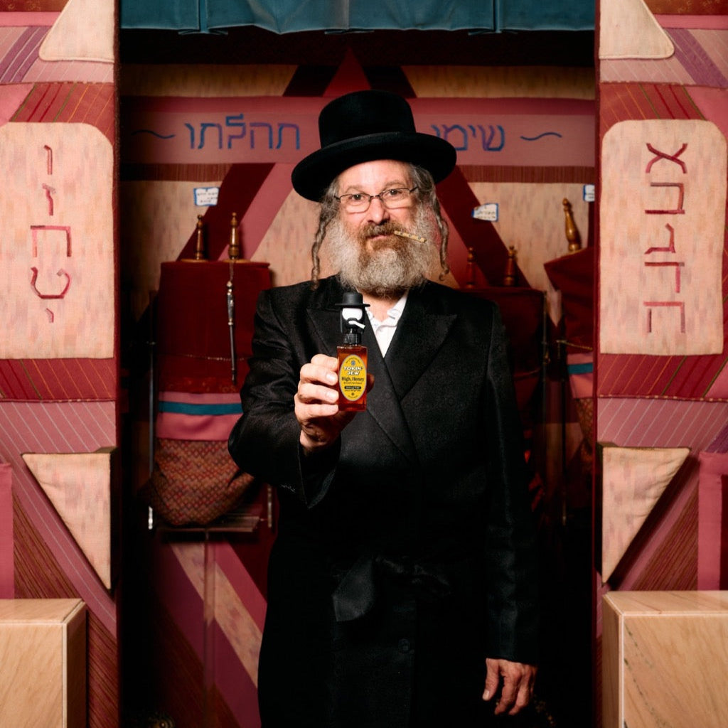 Rabbi in black coat and hat holding a honey bottle inside a decorated wooden structure with Hebrew text