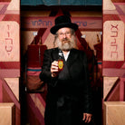 Rabbi in black coat and hat holding a honey bottle inside a decorated wooden structure with Hebrew text
