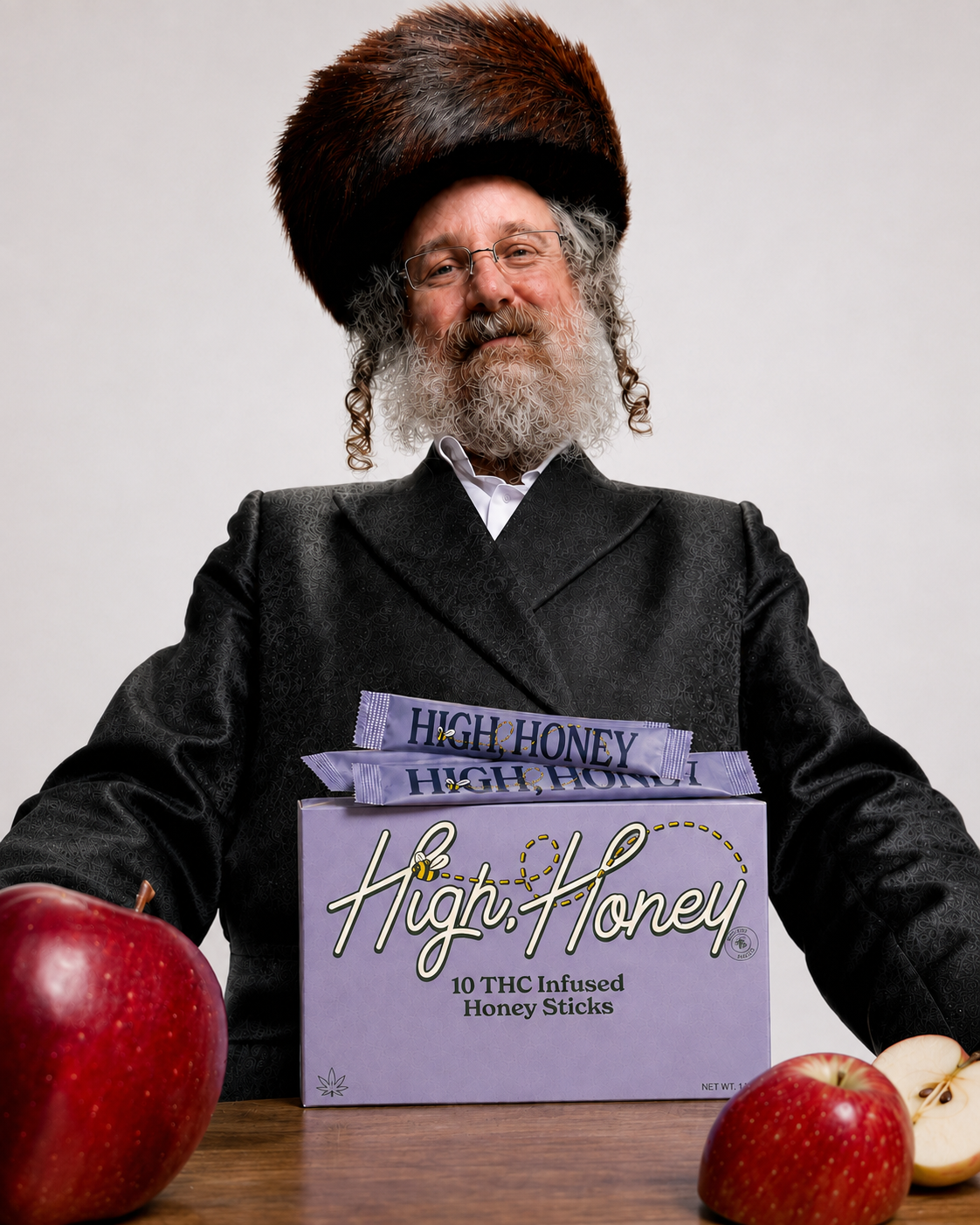A man wearing a dark shtreimel and black  coat sits behind a box of High Honey 10mg THC Infused Honey Sticks, with red apples on a table in the foreground against a light gray background.