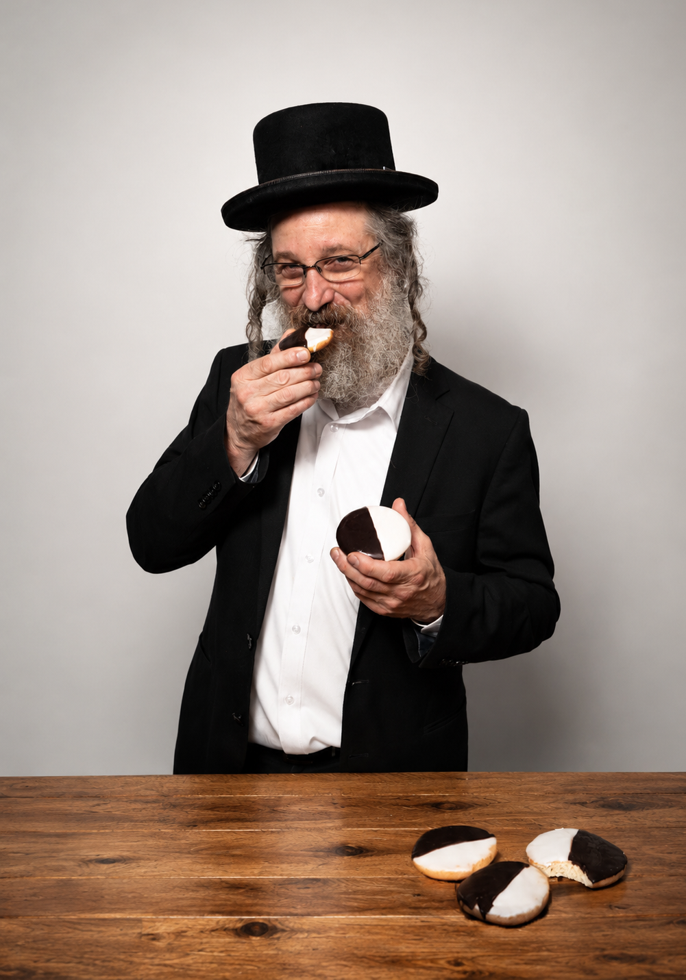 A man with a long gray beard and shtreimel, wearing a black suit and white shirt, eating a chocolate and white pastry while holding another in his hand, standing behind a wooden table with more pastries on a light gray background.