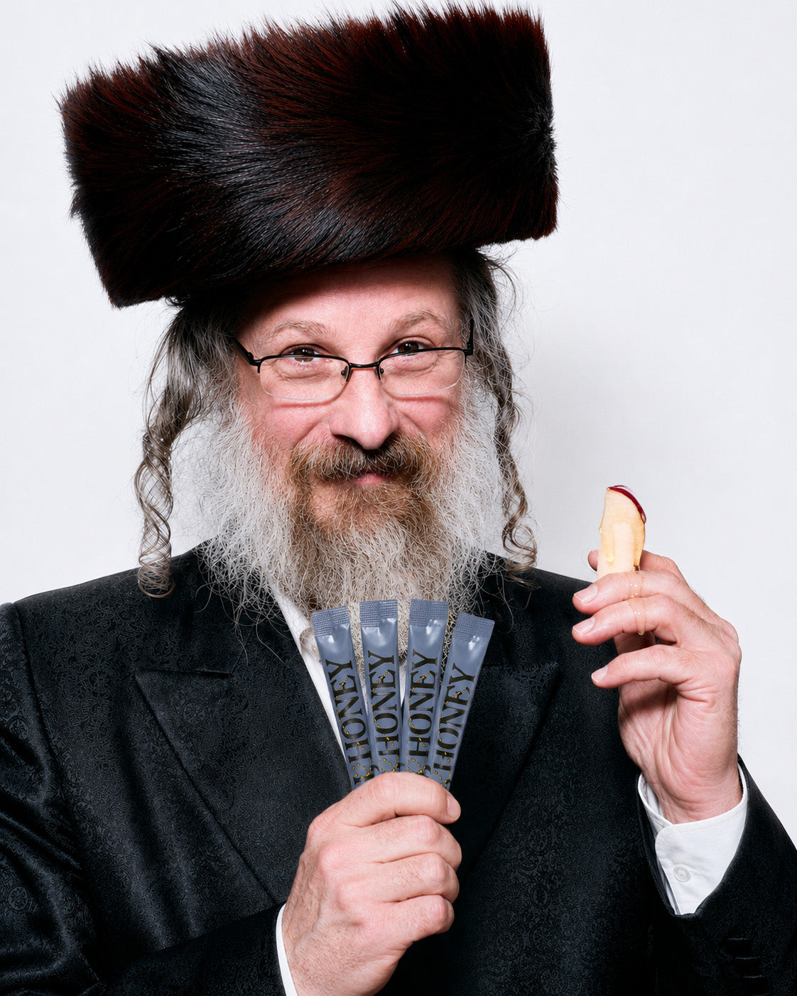 A Rabbi with a long gray beard and glasses, wearing a shtreimel and black suit jacket, holding four honey packets in one hand and a piece of apple in the other, against a plain light gray background.