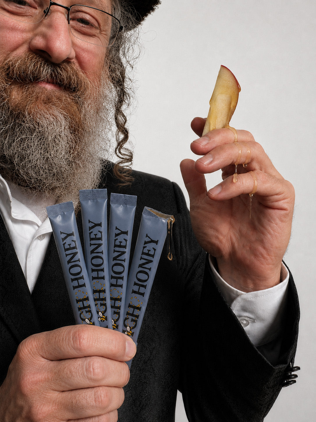 A bearded man in a black suit and shtreimel holds several blue honey sticks in one hand and a slice of apple with honey dripping from it in the other, against a plain light background.