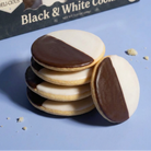 A box of Black & White Cookies sits behind a stack of four round cookies, with one cookie leaning in front. The cookies are frosted with dark brown and white icing in a half-and-half pattern, set on a light blue surface with a few crumbs.