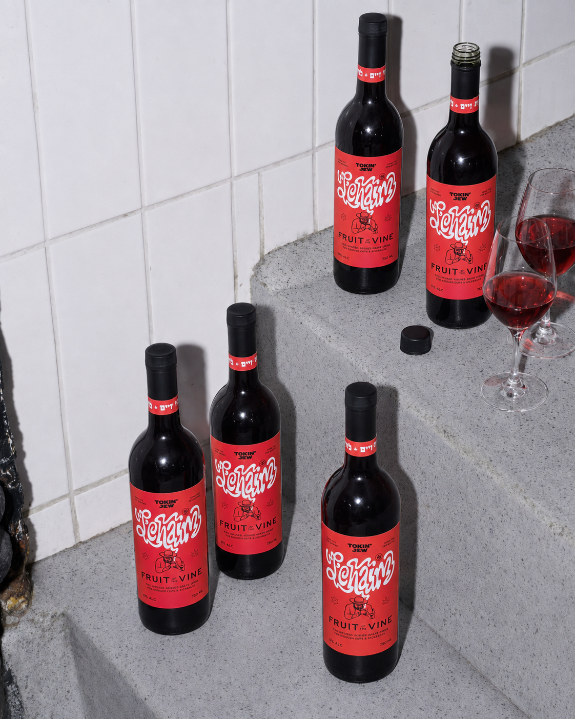 Four bottles of red wine with distinctive red and white labels are arranged on gray stone steps against a white tiled wall, with two filled wine glasses and a small black cap nearby.