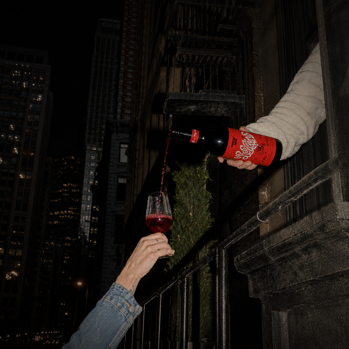 A person pours red juice from a bottle with a bold red label into a glass held by another person on a balcony at night, with city buildings in the background.